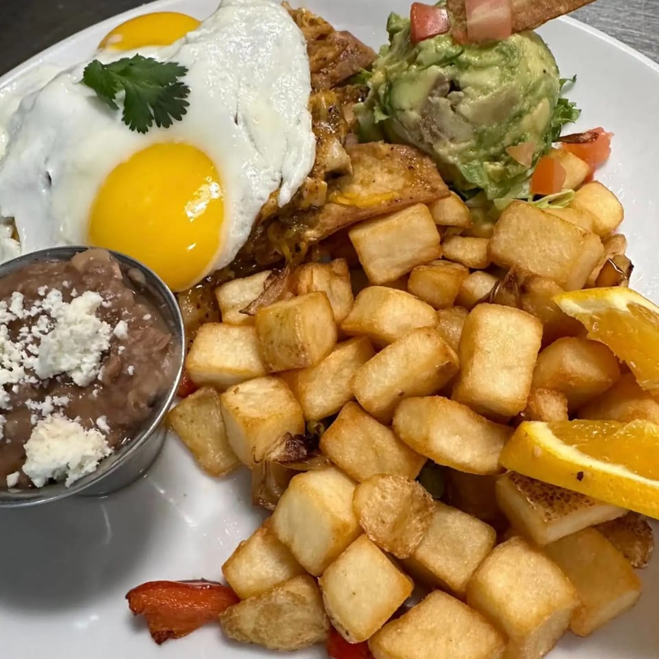 A breakfast plate with two fried eggs, home fries, refried beans, guacamole, and a tortilla chip.