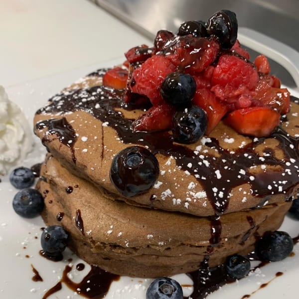 Stack of chocolate pancakes topped with fresh berries, chocolate syrup, and powdered sugar.