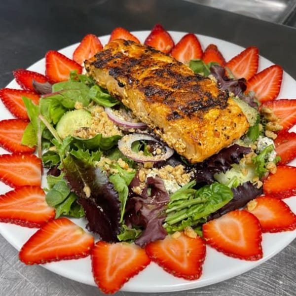 Grilled salmon fillet on a bed of mixed greens, surrounded by sliced strawberries.
