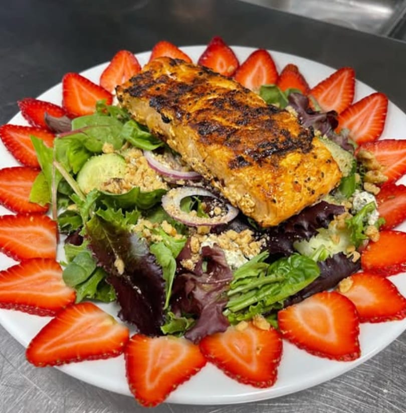 Grilled salmon fillet on a bed of mixed greens, surrounded by sliced strawberries.