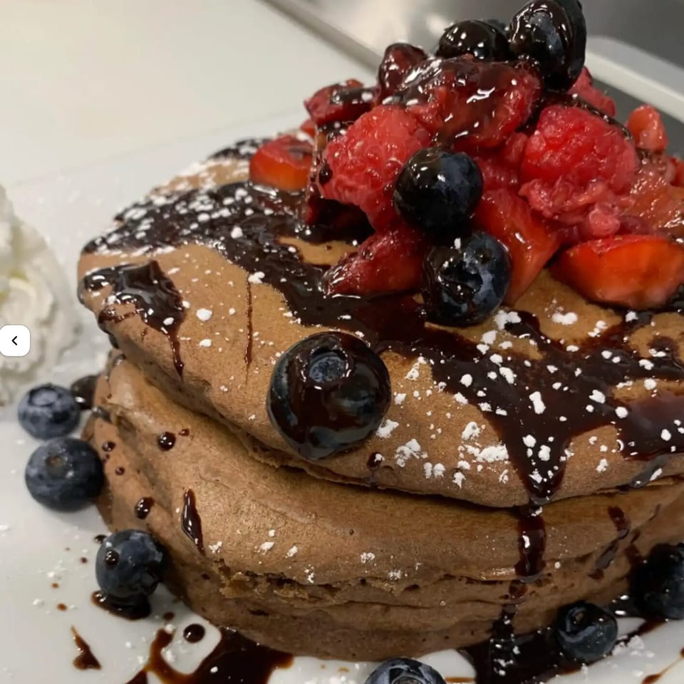 Stack of chocolate pancakes topped with fresh berries, chocolate syrup, and powdered sugar.